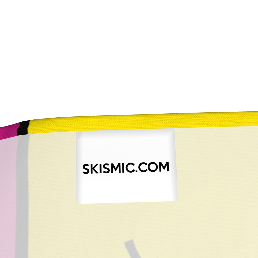 Close-up of a product edge showing a white label with the text 'SKISMIC.COM' in black, with a bright yellow trim and small pink and black accents against a pale cream background.